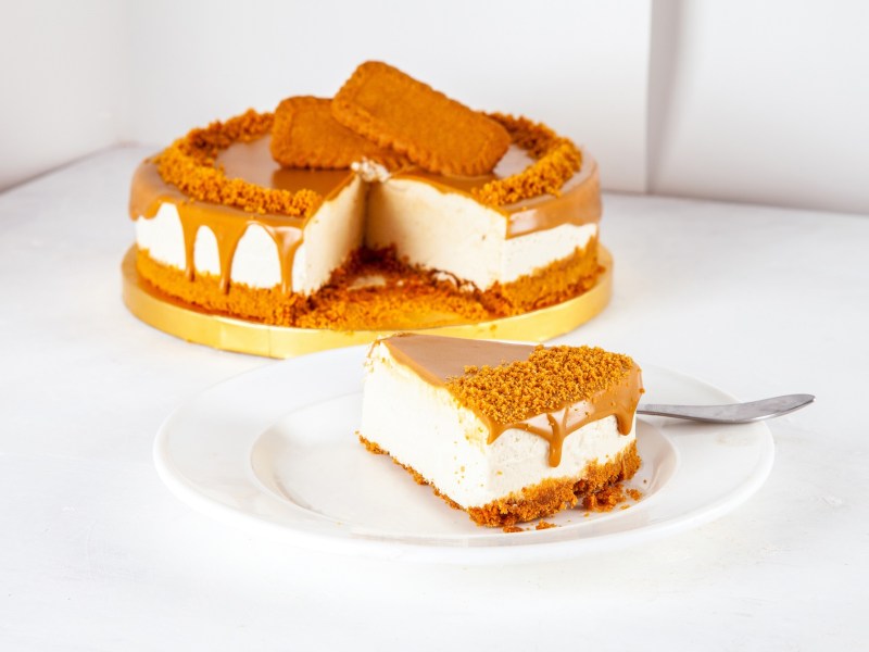 A slice of no-bake lotus biscoff cheesecake on a white plate. In the background is the rest of the cake on a yellow serving platter.