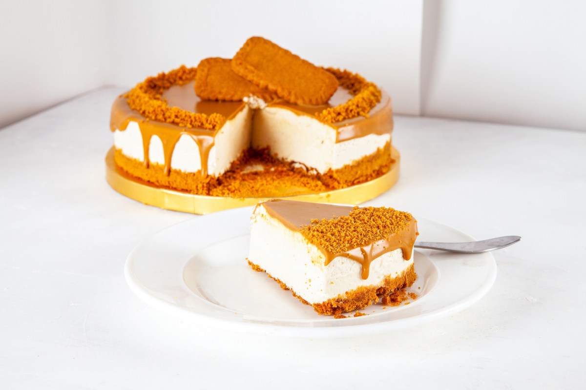 A slice of no-bake lotus biscoff cheesecake on a white plate. In the background is the rest of the cake on a yellow serving platter.