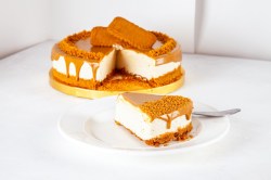 A slice of no-bake lotus biscoff cheesecake on a white plate. In the background is the rest of the cake on a yellow serving platter.