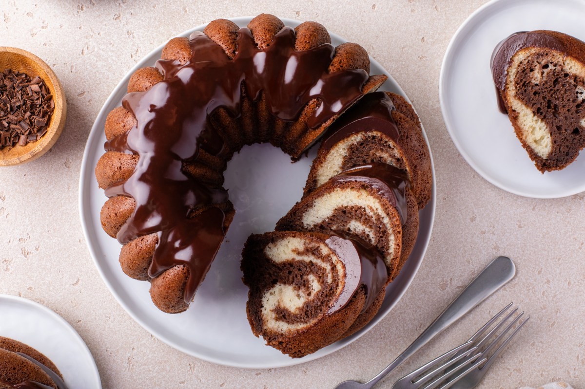 Top view of a sliced Nutella marble cake in a Bundt pan. The cake is on a white plate. Next to it on the right is another plate with a piece of marble cake with Nutella. To the left of it is a bowl of chocolate chips and there are some cake forks on the work surface.