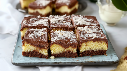 A large rectangular plate with square pieces of chocolate coconut cake topped with a chocolate glaze and shredded coconut. Behind the plate you can see another plate with chocolate coconut cake pieces. You can also see a white cloth lying around. And a glass container with milk or cream can be seen.