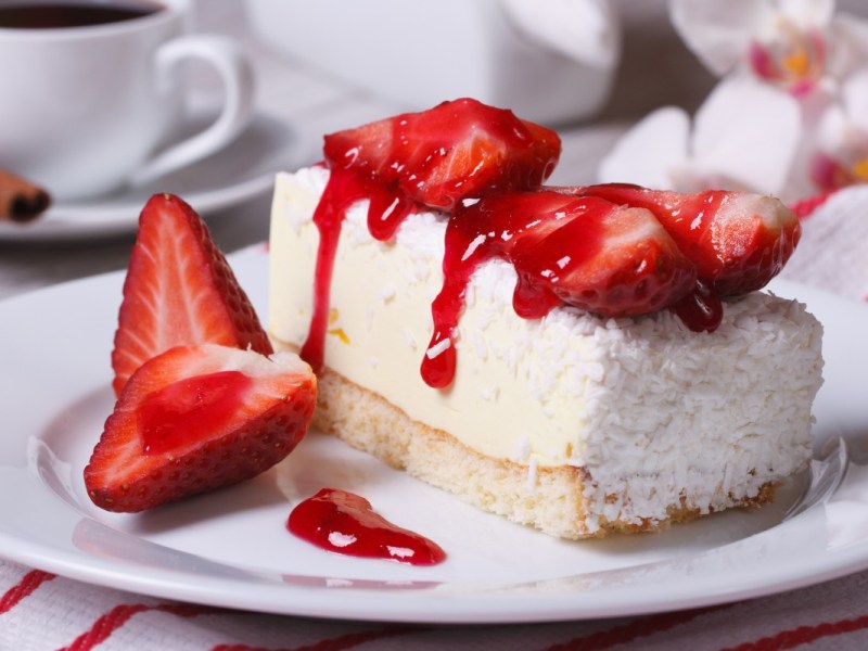 A piece of strawberry coconut cake on a plate. A cup of coffee can be seen in the background.