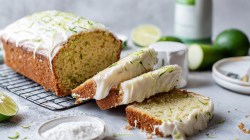 A sliced sweet zucchini cake with white cream cheese frosting. Surrounding it are ingredients such as fresh limes, a bowl of powdered sugar, and a sliced zucchini, as well as other containers and a plate.