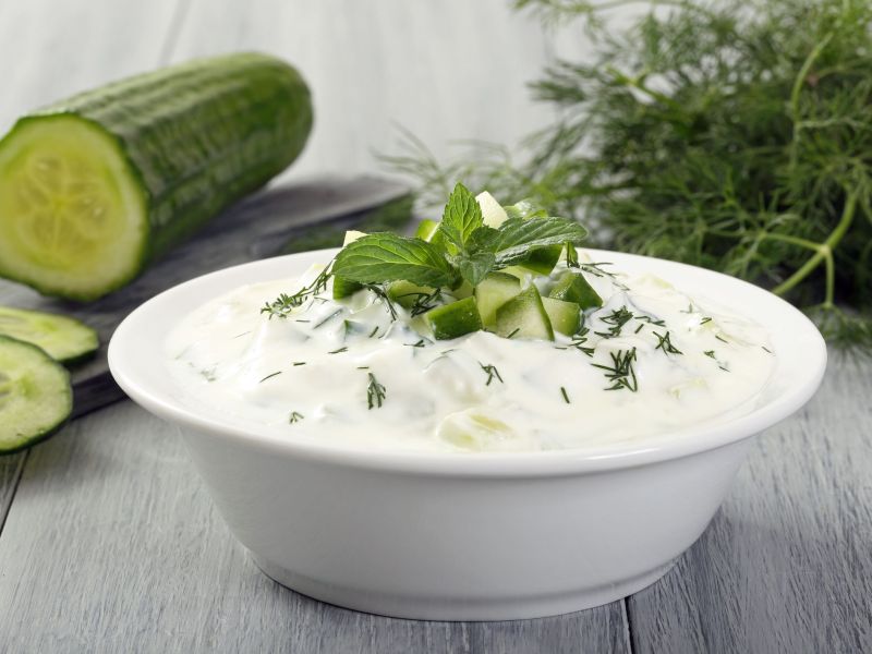 Tzatziki in a bowl, garnished with fresh herbs. In the background lies a sliced cucumber and more fresh herbs.