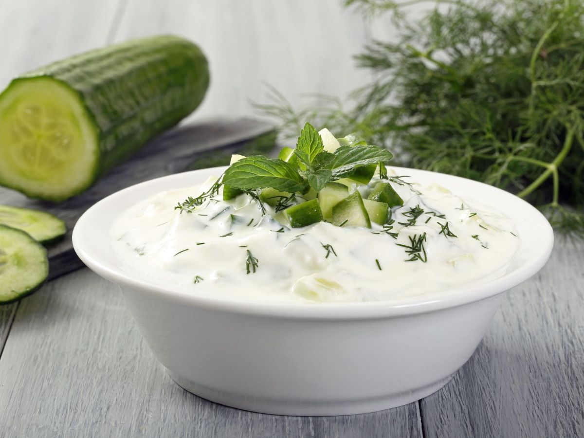 Tzatziki in a bowl, garnished with fresh herbs. In the background lies a sliced cucumber and more fresh herbs.