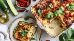 Overhead view of a baking dish with zucchini-feta lasagna. A piece of it lies on a plate next to it. Fresh ingredients such as zucchini, a bowl of sun-dried tomatoes, a glass jar with olive oil, fresh basil are scattered around.