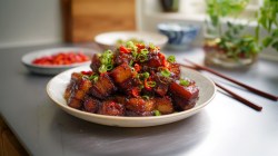 A plate of Coca-Cola glazed pork belly atop a plate with chopped green onions and chilies in full frontal view with bright white lighting.