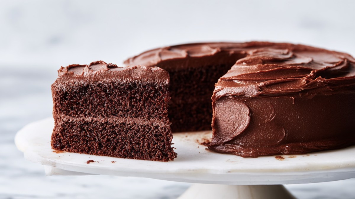 A cut chocolate cake with simple chocolate frosting on a cake stand in frontal view with white lighting.