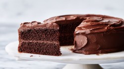 A cut chocolate cake with simple chocolate frosting on a cake stand in frontal view with white lighting.
